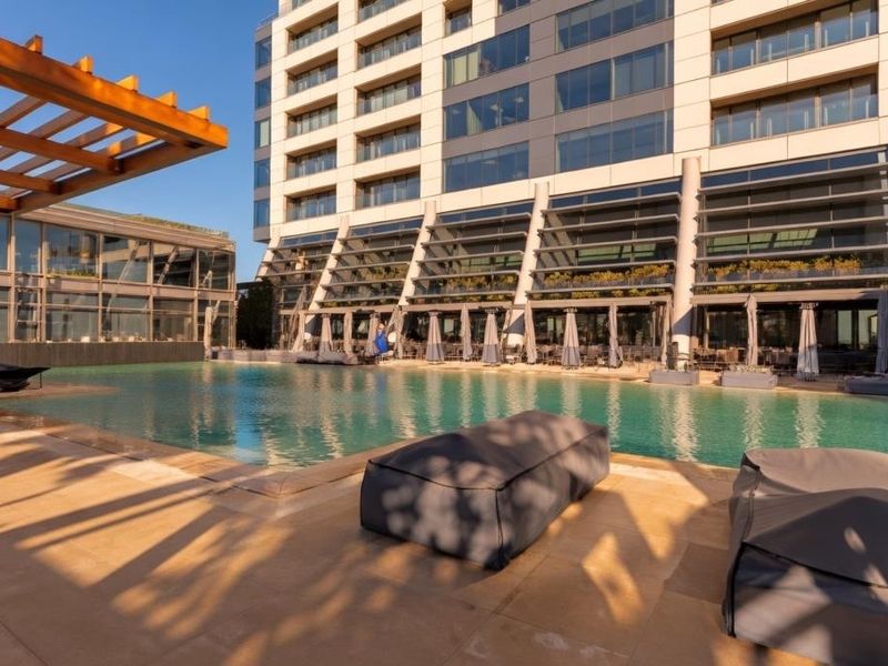 A modern outdoor swimming pool area featuring a timber pergola and paved decking, set against a contemporary high-rise apartment building facade.