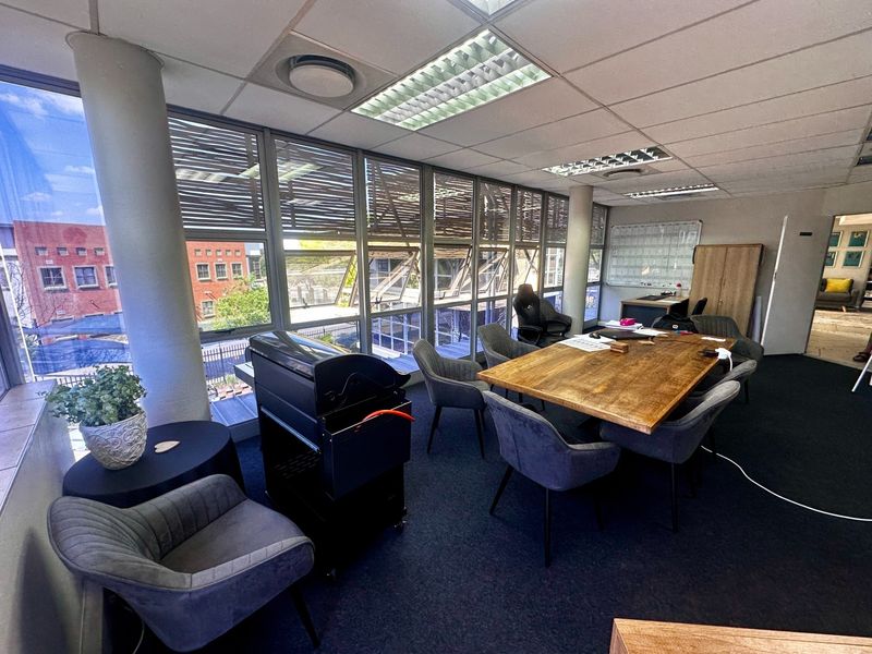A bright office space featuring large floor-to-ceiling windows, recessed lighting, and carpeted flooring, offering a professional work environment with natural light.