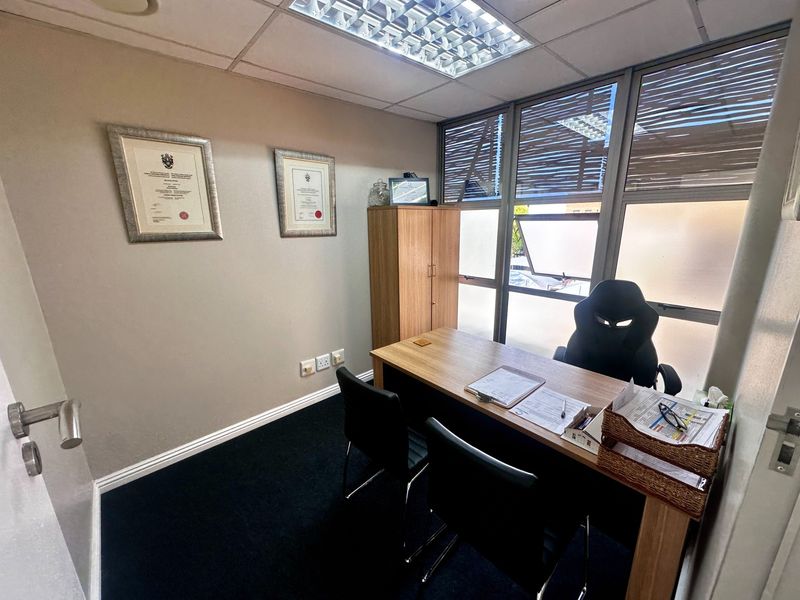 A functional office space featuring carpeted flooring, a suspended ceiling with integrated lighting, and large aluminium-framed windows providing natural light.