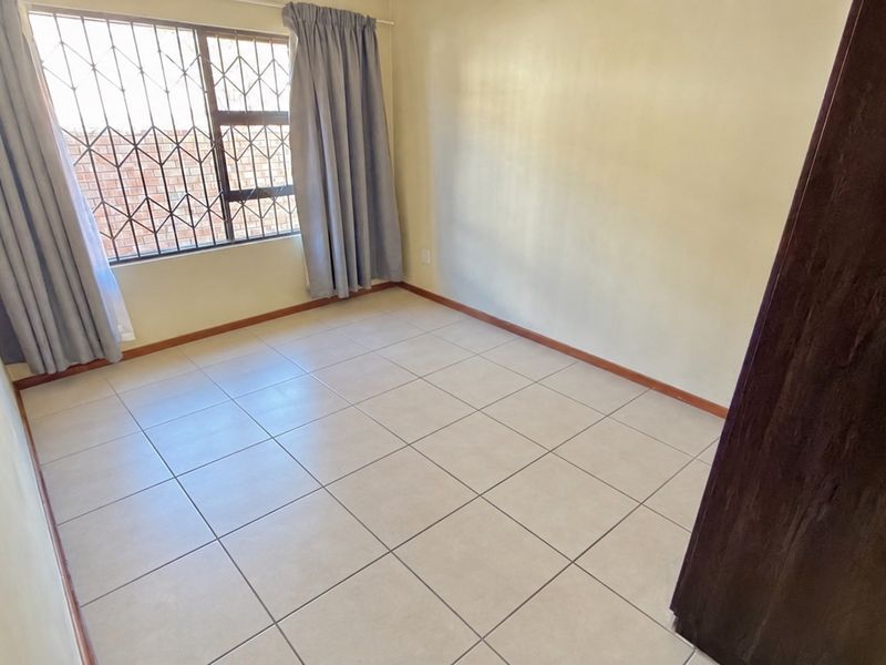 A clean, empty bedroom featuring neutral tiled flooring, a built-in cupboard, and a window fitted with security bars.