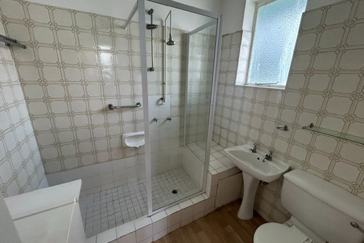A functional bathroom featuring a walk-in shower with glass enclosure, pedestal basin, and toilet, finished with dated wall tiling and a small window.