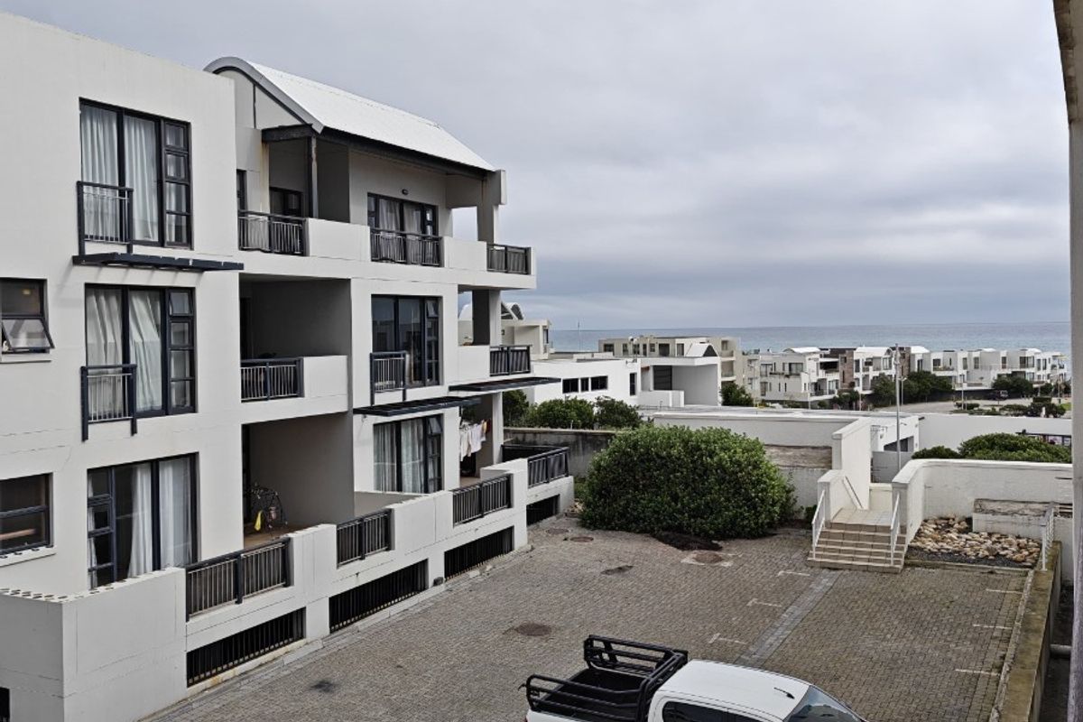 Exterior view of the apartment complex rear facade, featuring paved parking area, multiple balconies, and coastal neighborhood surroundings.