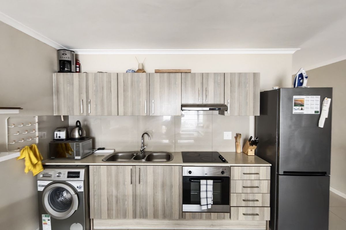 A functional kitchen featuring light wood-grain cabinetry, integrated electric hob and oven, stainless steel sink, and tiled splashback with neutral floor tiles.