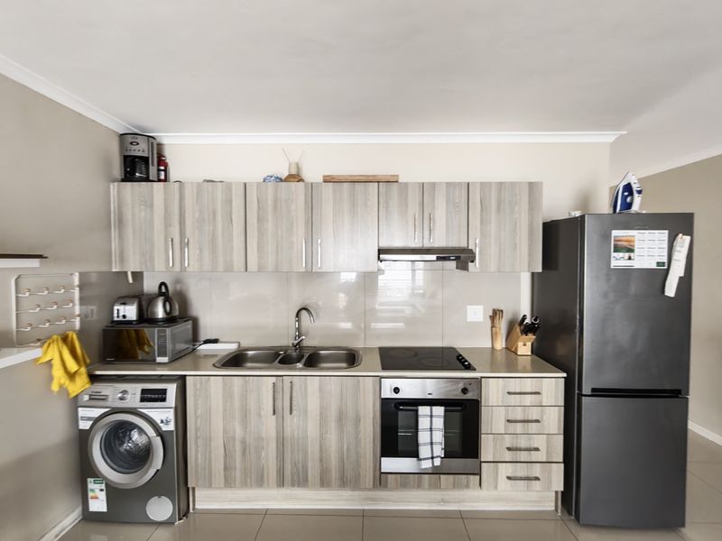 A functional kitchen featuring light wood-grain cabinetry, integrated electric hob and oven, stainless steel sink, and tiled splashback with neutral floor tiles.