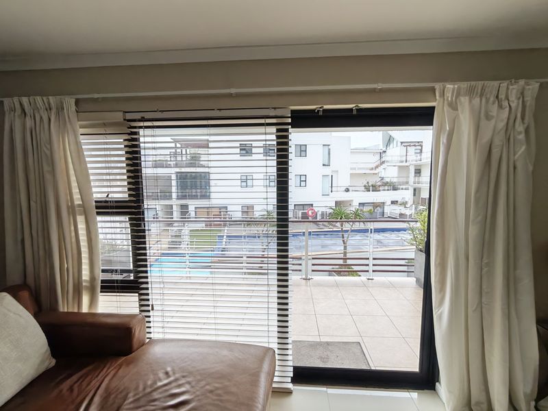Living area featuring a large sliding glass door with venetian blinds and curtains, providing direct access to a tiled balcony overlooking the complex pool.