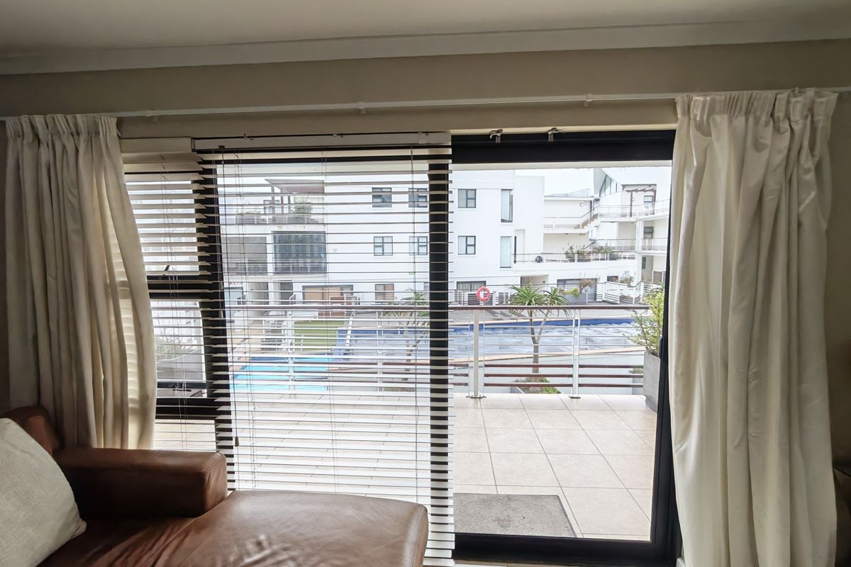 Living area featuring a large sliding glass door with venetian blinds and curtains, providing direct access to a tiled balcony overlooking the complex pool.