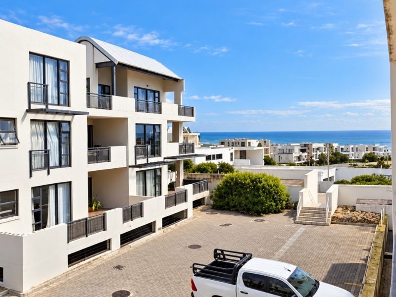 Exterior view of the apartment complex featuring paved parking area, balconies with metal railings, and a scenic coastal backdrop.