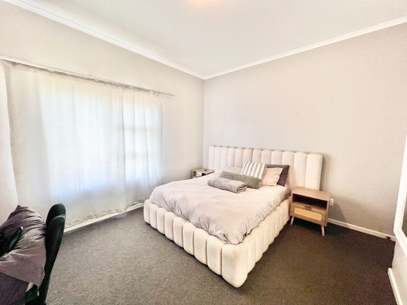 A well-maintained bedroom featuring neutral wall tones, dark carpeting, and a large window with a curtain rod.