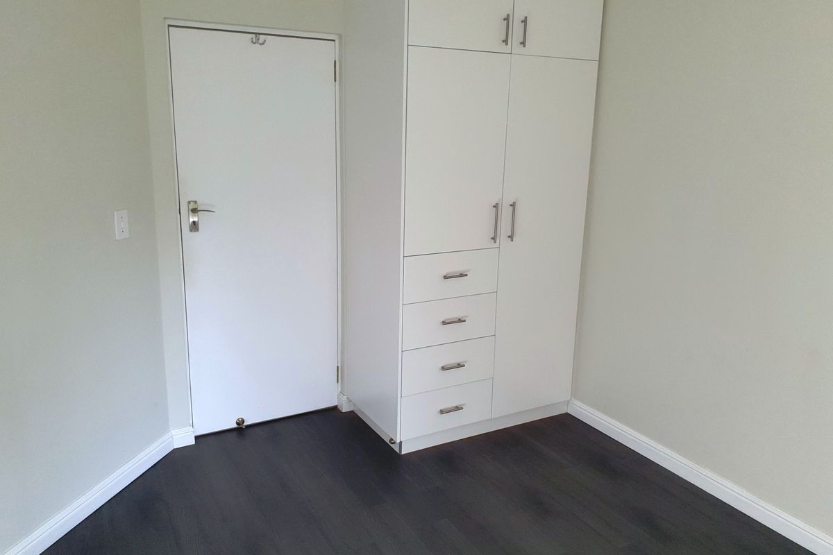 A clean, empty bedroom featuring dark laminate flooring, neutral walls, and a white built-in wardrobe with integrated drawers.