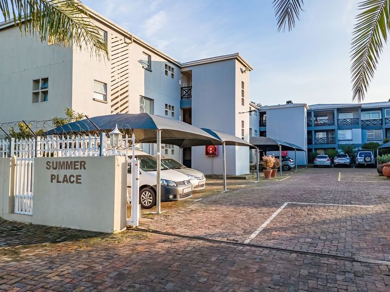 Front exterior view of Summer Place apartment complex featuring a paved driveway, covered parking bays, and secure perimeter fencing.
