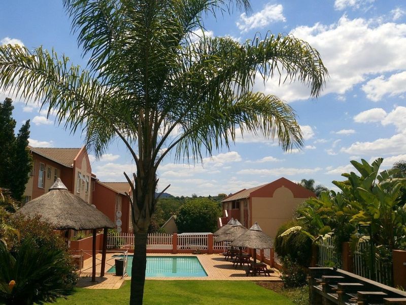 Communal pool area featuring a swimming pool, paved deck, and thatched lapa structures within a well-maintained apartment complex garden setting.