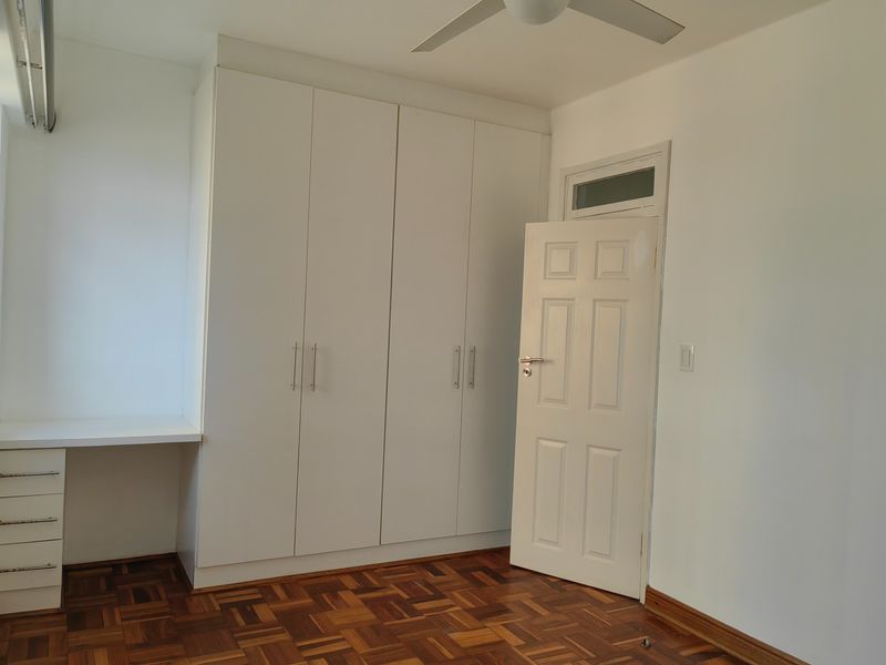 A bright bedroom featuring classic parquet flooring, ample built-in cupboard storage, a dedicated built-in desk area, and a ceiling fan.