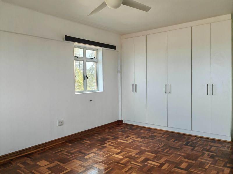 A bright, empty bedroom featuring classic parquet flooring, ample built-in cupboard storage, and a ceiling fan.