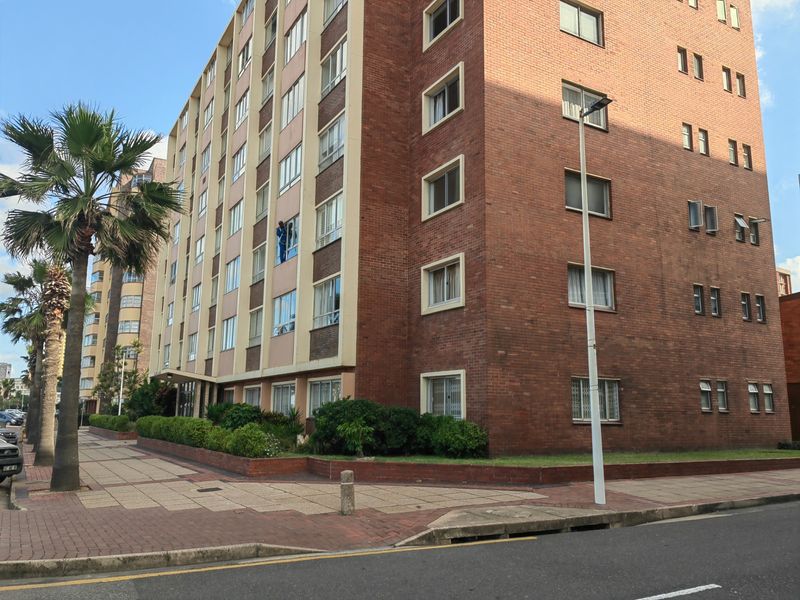 Exterior view of a multi-story brick apartment building featuring paved walkways and established garden beds along the street frontage.