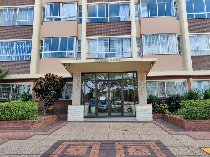 The front entrance of the Dunelands apartment complex, featuring a glass door, brick facade, and paved walkway with established garden beds.