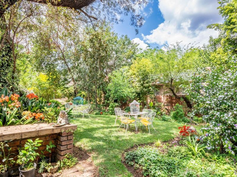 A lush, well-maintained garden featuring mature trees, vibrant flowering plants, and a rustic brick retaining wall, offering a serene outdoor space.