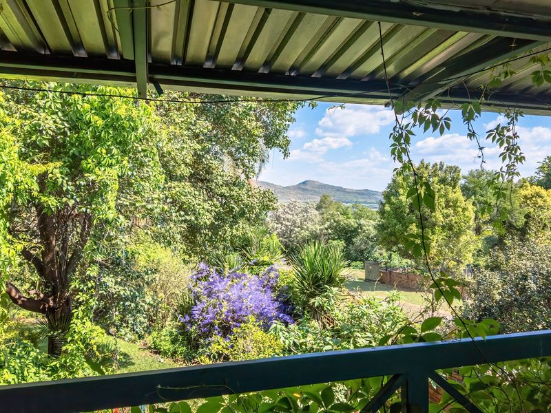 A covered balcony area featuring a metal roof and railing, offering an elevated view over a lush, mature garden and distant hills.