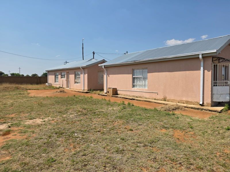 Rear view of the property showing two attached residential units with plastered walls, corrugated metal roofing, and security gates set on a large plot.