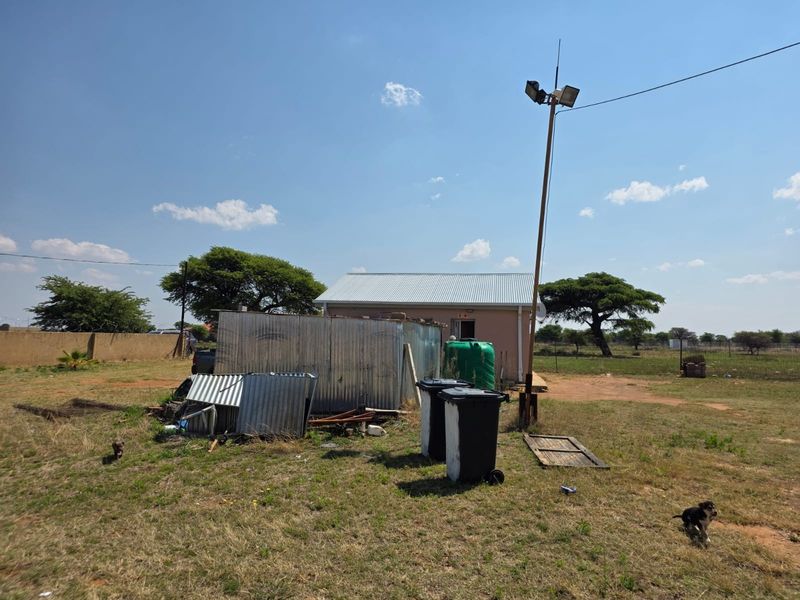 Rear view of the small holding property featuring a corrugated iron utility structure, water tank, and floodlight pole on a large open plot.
