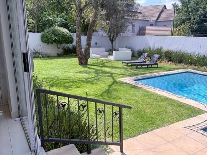 A well-maintained backyard featuring a swimming pool with a safety net, paved patio area, and lush green lawn enclosed by a perimeter wall.