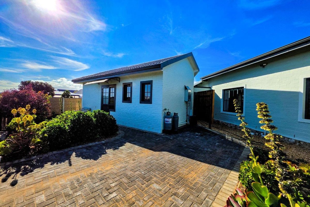 A well-maintained rear exterior view featuring a paved courtyard, painted brick walls, and established garden beds under a clear blue sky.