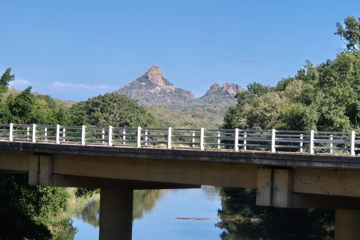 A scenic view featuring a concrete bridge spanning a calm river with prominent mountain peaks in the background.