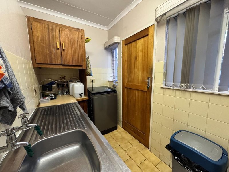 A functional laundry room featuring a stainless steel sink, tiled splashbacks, wooden cabinetry, and direct access via a solid wood door.