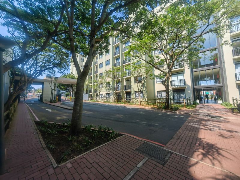 Exterior view of the apartment complex entrance featuring paved driveway, mature trees, and multi-story building facade with balconies.