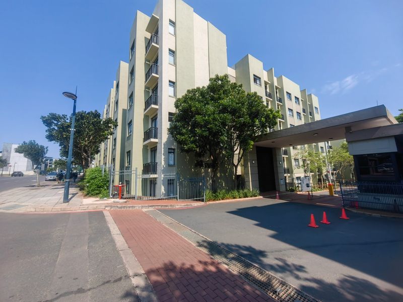 Exterior view of a modern apartment complex featuring a secure gated entrance, paved driveway, and multi-story building with private balconies.