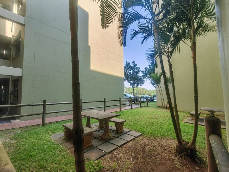 A communal garden area between apartment blocks featuring a paved walkway, timber fencing, and a concrete outdoor seating area.