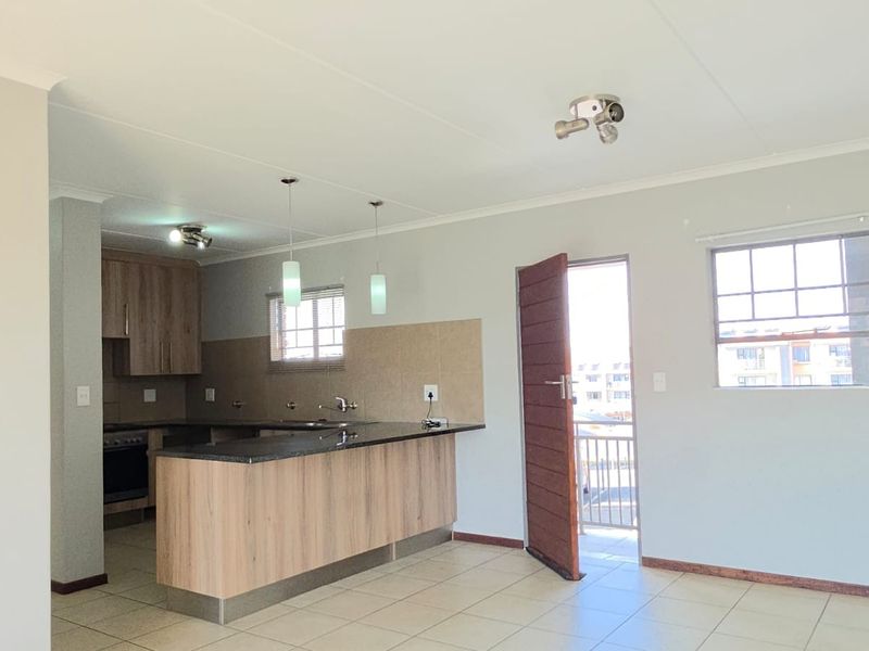 Open-plan living area featuring a modern kitchen with wood-finish cabinetry, granite countertops, and tiled flooring, leading to an exterior balcony.
