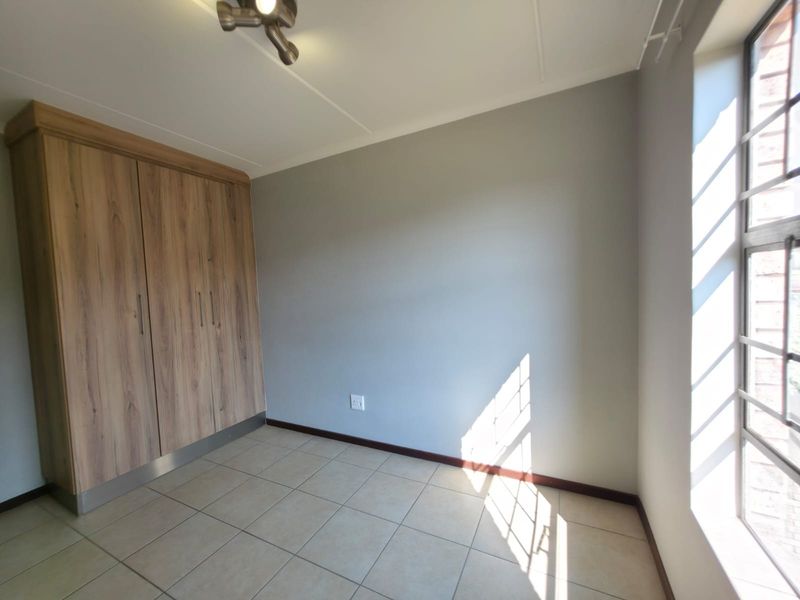 A bright, empty bedroom featuring neutral tiled flooring, built-in wooden cupboards, and large windows with security bars.