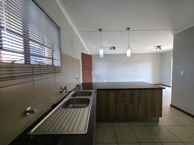 An open-plan kitchen area featuring wood veneer cabinetry, granite countertops, a double sink, and tiled flooring, flowing into an adjacent living space.