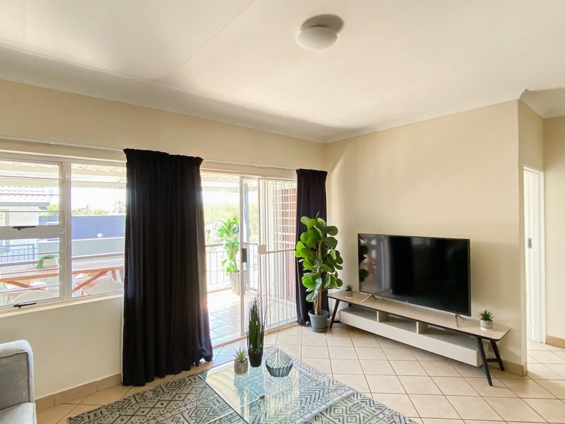 A bright living room featuring tiled flooring, a sliding glass door leading to a balcony, and large windows with security bars.