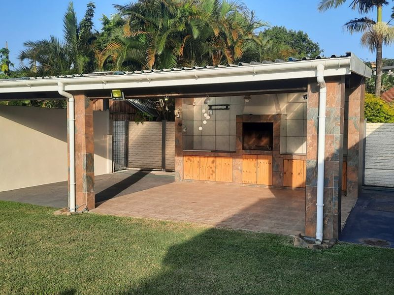 A spacious covered outdoor patio featuring a built-in braai, tiled flooring, and brick pillars, overlooking a well-maintained garden area.