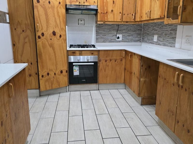 A clean, functional kitchen featuring knotty pine cabinetry, stone countertops, a gas hob with extractor fan, and modern light-toned tiled flooring.