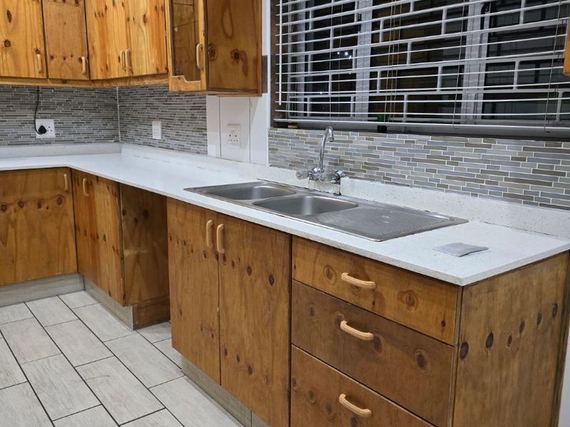 A functional kitchen featuring wooden cabinetry, stone countertops, a double basin sink, and modern mosaic tile backsplash with light-colored ceramic floor tiles.