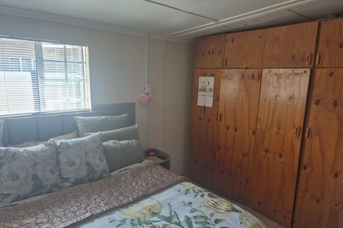 A bedroom featuring built-in wooden cupboards, an aluminium window with blinds, and exposed ceiling and electrical finishes.