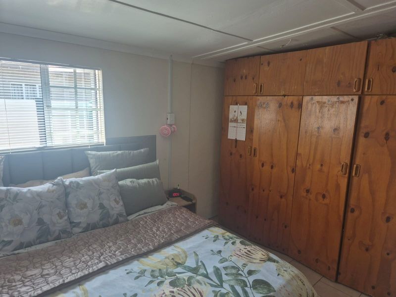 A bedroom featuring built-in wooden cupboards, an aluminium window with blinds, and exposed ceiling and electrical finishes.
