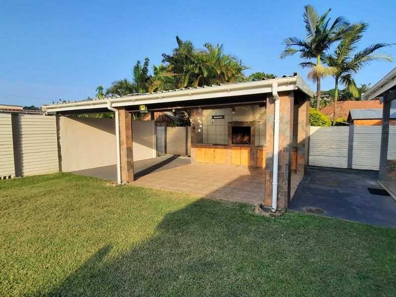A spacious covered patio featuring a built-in braai, stone-clad pillars, and tiled flooring, overlooking a well-maintained garden area.
