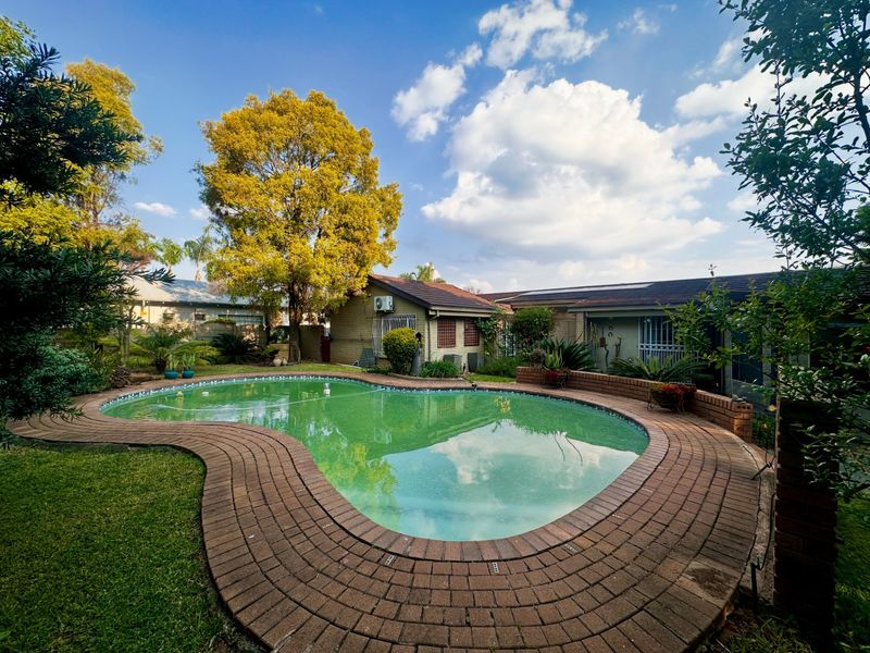 Outdoor pool area featuring a kidney-shaped swimming pool with brick surround and established garden landscaping.