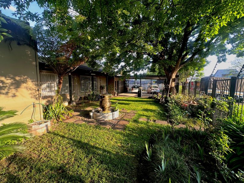 A lush, established garden featuring a central stone fountain, mature trees, paved pathways, and secure perimeter fencing with an attached carport.