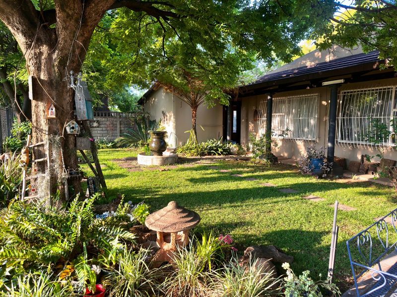 A lush, well-maintained garden featuring mature trees, a central stone fountain, and a paved area leading to the home's exterior facade.