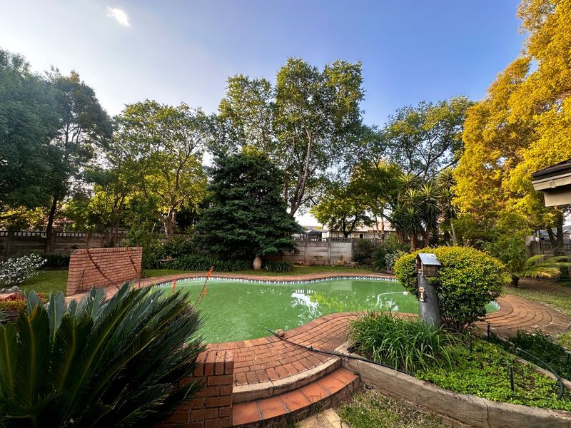 A backyard swimming pool area featuring brick paving, a retaining wall, and mature garden landscaping.