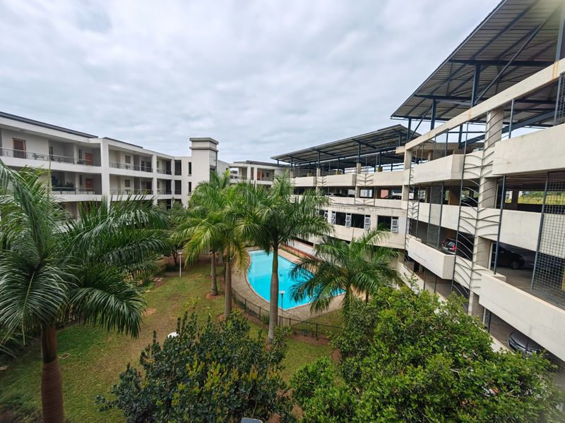 An elevated view of a residential apartment complex featuring a central swimming pool, landscaped garden area, and a multi-level parking structure.