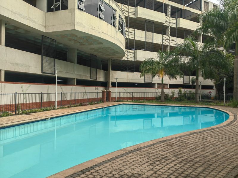 A communal swimming pool area featuring paved surrounds and metal fencing, set against the backdrop of a multi-level concrete apartment building.