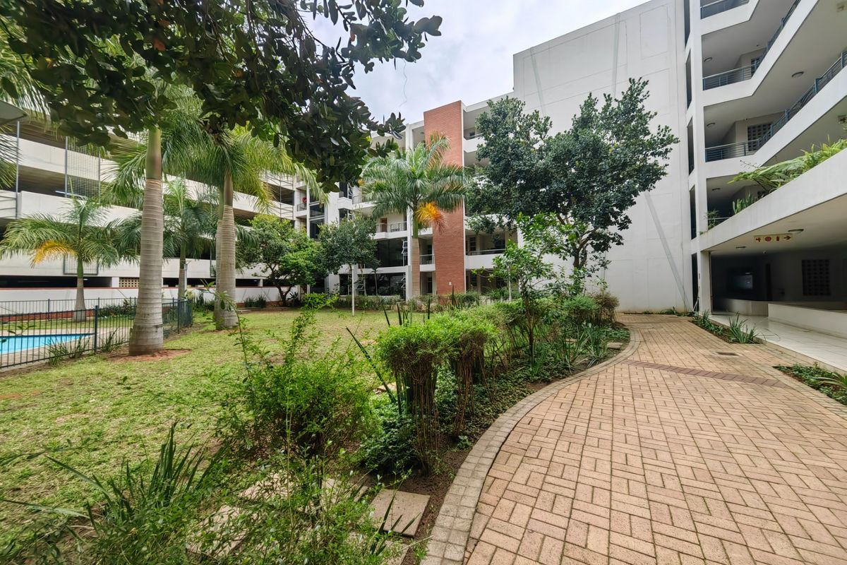 A view of the apartment complex communal garden area featuring a paved walkway, landscaped greenery, and access to the swimming pool.