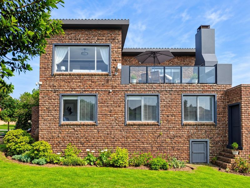 Rear elevation of a modern double-story brick house featuring a spacious balcony with glass balustrades, an outdoor fireplace, and well-maintained garden landscaping.
