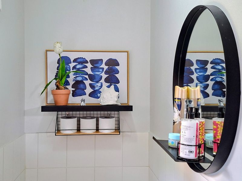 A close-up view of a bathroom featuring a modern black circular mirror and matching wall-mounted storage shelving above white ceramic wall tiles.