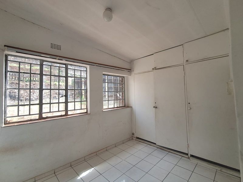 An empty bedroom featuring tiled flooring, built-in cupboards, and steel-framed windows with security bars, requiring cosmetic updates to improve its condition.